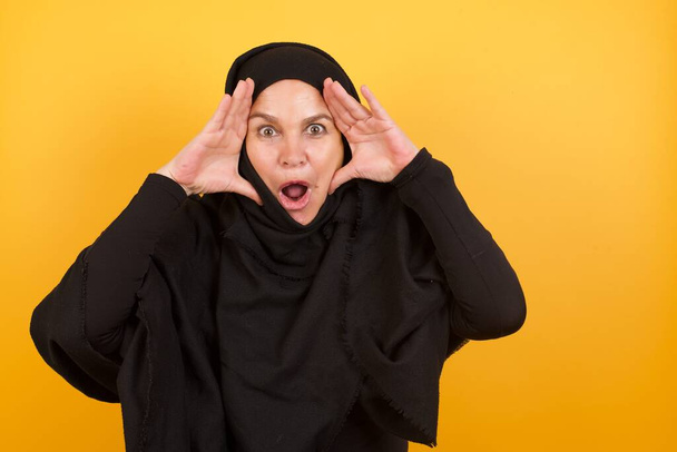 Donna matura che indossa hijab con espressione spaventata, tiene le mani sulla testa, mascella caduta, ha un'espressione formidabile. Concetto di mg - Foto, immagini