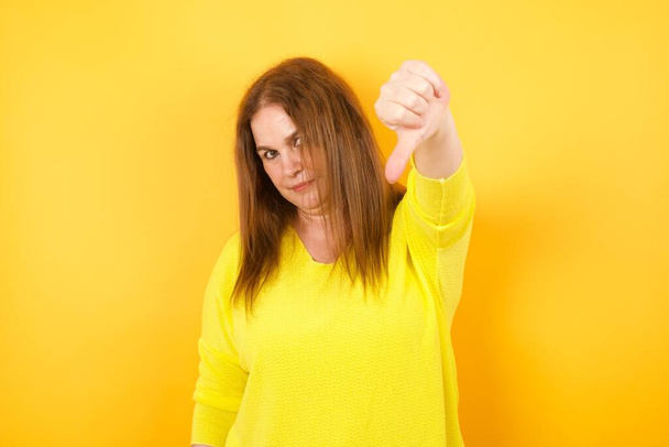 νεαρή γυναίκα αισθάνεται θυμωμένη, ενοχλημένη, απογοητευμένη ή δυσαρεστημένη, δείχνοντας τους αντίχειρες κάτω με ένα σοβαρό βλέμμα ενάντια στο λευκό τοίχο - Φωτογραφία, εικόνα