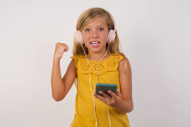 cute little girl posiada nowoczesny telefon komórkowy podłączony do słuchawek, zaciska pięść od dobrych emocji, wykrzykuje z radością,  - Zdjęcie, obraz
