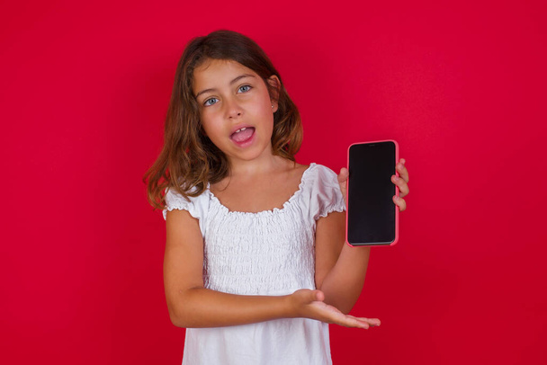 Cute little girl with smartphone posing on red studio background - Фото, зображення