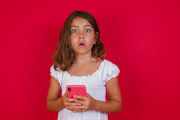 Симпатична маленька дівчинка зі смартфоном позує на червоному фоні студії
 - Фото, зображення
