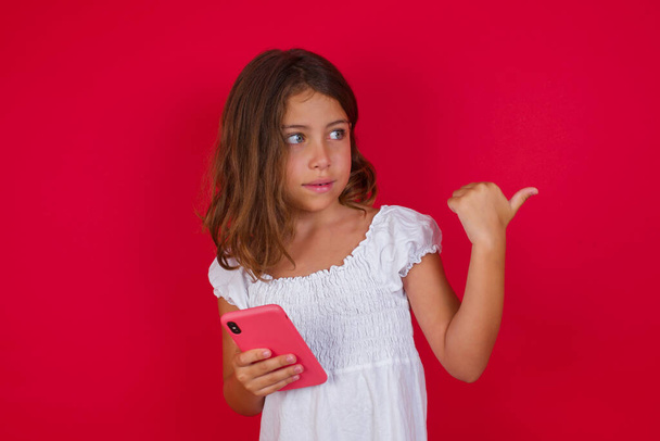 Schattig klein meisje met smartphone poseren op rode studio achtergrond - Foto, afbeelding