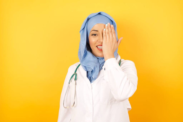 La joven doctora vistiendo uniforme médico cubriendo un ojo con su mano tiene una sonrisa confiada. Concepto de salud. - Foto, imagen