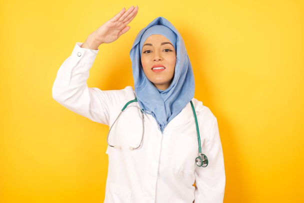 Όμορφη γυναίκα Άραβας γιατρός με στηθοσκόπιο κρατώντας το χέρι κοντά στο μέτωπο πάνω από κίτρινο φόντο στούντιο - Φωτογραφία, εικόνα