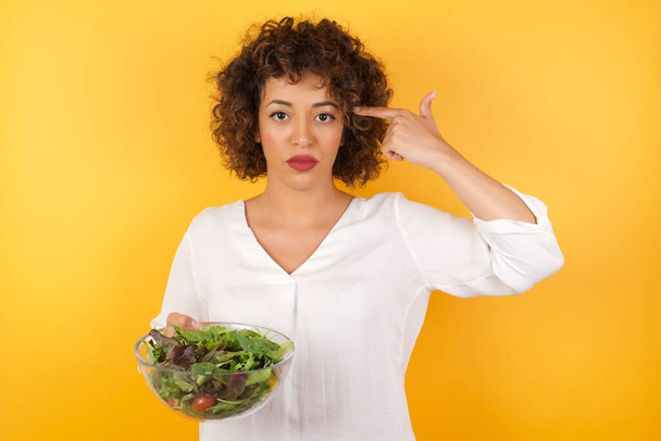 Caucasian woman with curly hair in studio with yellow wall, holding glass bowl with fresh salad food, suicide gesture, gunshot at head  - Photo, Image
