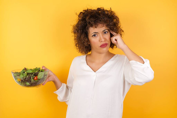 Beauitul woman holding bowl with salad,  confused and annoyed with open palm showing copy space and pointing finger to forehead. Think about it. - Photo, Image
