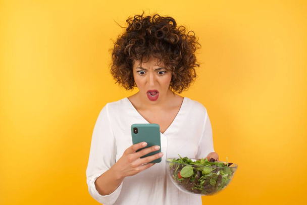 Portrait de femme choquée regardant un smartphone, lisant les nouvelles des médias sociaux, ou e-mail important porte chemise rouge, debout sur fond jaune. - Photo, image