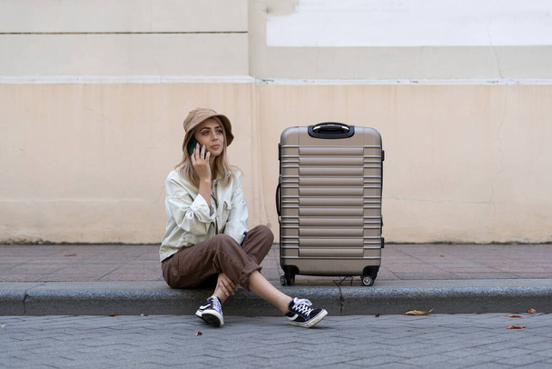 turistka s kufrem v evropském městě, cestovní ruch v Evropě. Sedí na chodníku a mluví po telefonu. Místo pro text. Kopírovat prostor. - Fotografie, Obrázek