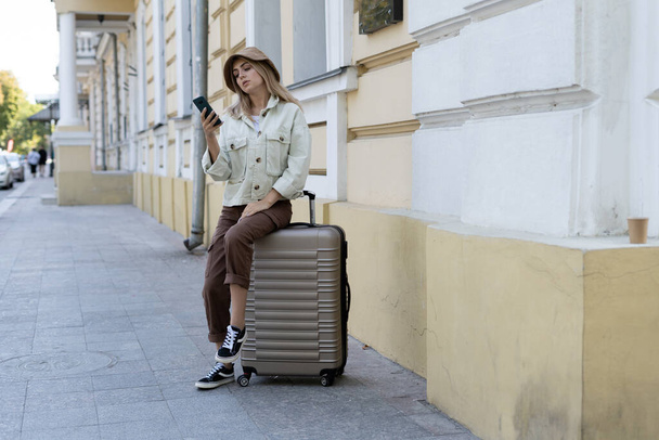 tourist woman in a European city, tourism in Europe. Sits on a large suitcase on the sidewalk typing on the phone. Copy space. - 写真・画像