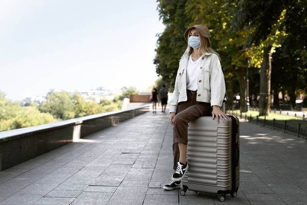 ヨーロッパの都市で観光客の女性、ヨーロッパの観光。医療保護マスクを着た少女がスーツケースの上に座っている。スペースのコピー. - 写真・画像