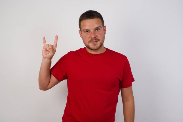 Νεαρός Καυκάσιος κάνει μια ροκ χειρονομία και χαμογελά στην κάμερα. Φορώντας κόκκινο πουκάμισο στέκεται πάνω σε λευκό τοίχο - Φωτογραφία, εικόνα