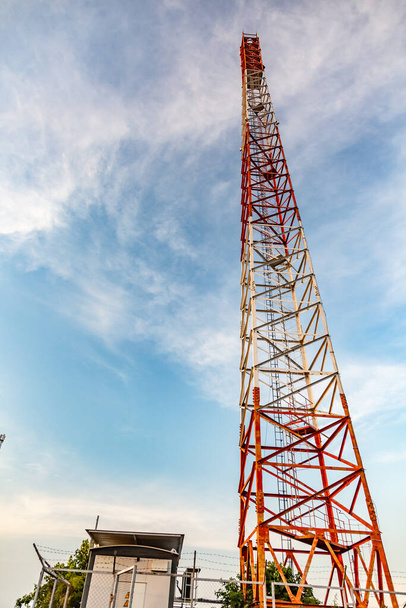 Una torre metálica para la comunicación móvil en rojo y blanco. Antena y pequeños edificios cercanos contra el cielo azul. Orientación vertical. Foto de alta calidad - Foto, Imagen