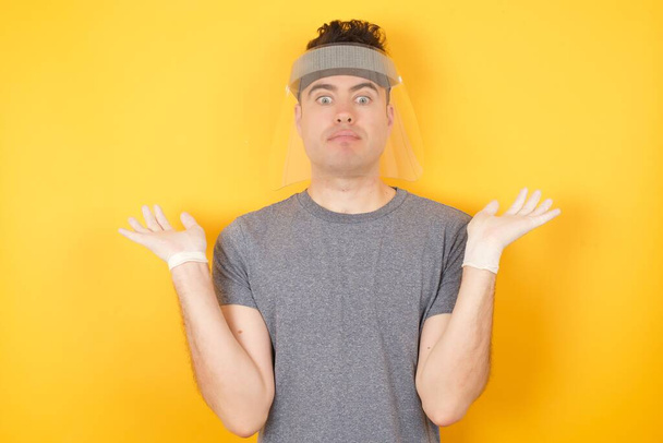 mężczyzna w plastikowym kapeluszu ochronnym pozowanie w studio na żółtym tle, wzrusza ramionami, wygląda niepewnie i zdezorientowany  - Zdjęcie, obraz