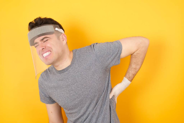 Junger kaukasischer Mann mit T-Shirt und Gesichtsschutz, der vor isoliertem gelben Hintergrund steht, leidet unter Rückenschmerzen, Berührung mit der Hand, Muskelschmerzen - Foto, Bild