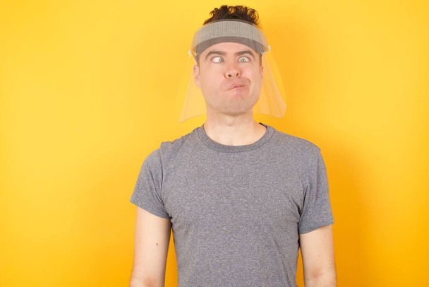 Young caucasian man wearing t-shirt and face shield standing over isolated yellow background making grimace and crazy face, screaming out of control, funny lunatic expressing freedom and wild. - Photo, Image