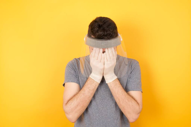 Young caucasian man wearing t-shirt and face shield standing over isolated yellow background covering her face with her hands, being devastated and crying. - Photo, Image
