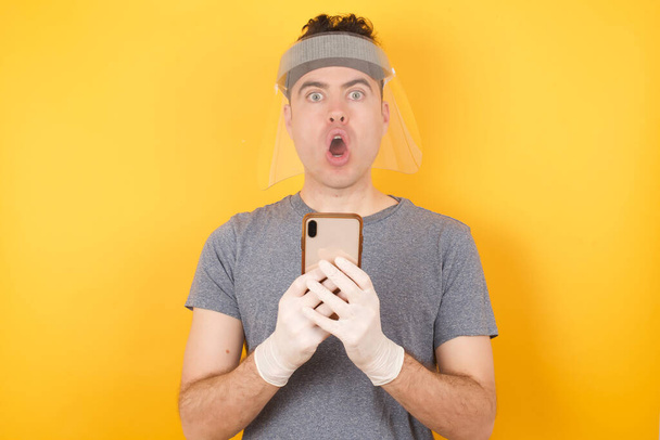 Молодий білий чоловік у футболці та щиті, що стоїть над ізольованим жовтим тлом, відкриває рот, тримає телефон, читаючи рекламу, неймовірні великі ціни на покупки
  - Фото, зображення