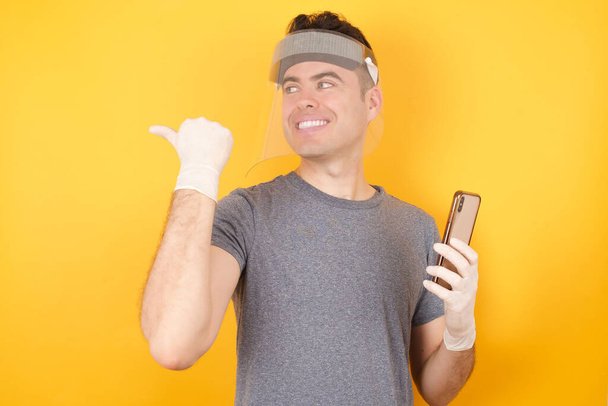 Молодой кавказский мужчина в футболке и защитном щите стоит на изолированном желтом фоне, используя и смс со смартфоном на изолированном фоне, указывая и показывая с большим пальцем в сторону со счастливым лицом, улыбающимся - Фото, изображение