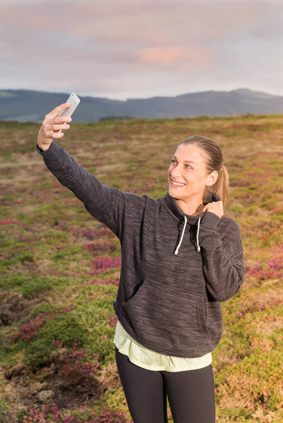 Spor kıyafetli kız gün batımında parkta cep telefonuyla kendi portresini çekiyor. - Fotoğraf, Görsel