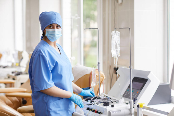 Retrato de enfermeira mascarada e de uniforme olhando para a câmera enquanto trabalhava com equipamentos médicos para doação de sangue no hospital - Foto, Imagem