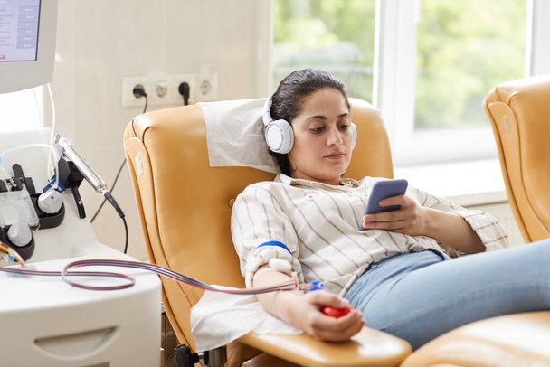Fiatal nő a fülhallgatóban mobiltelefonnal zenét hallgat, miközben fekszik a kanapén és vért adományoz. - Fotó, kép