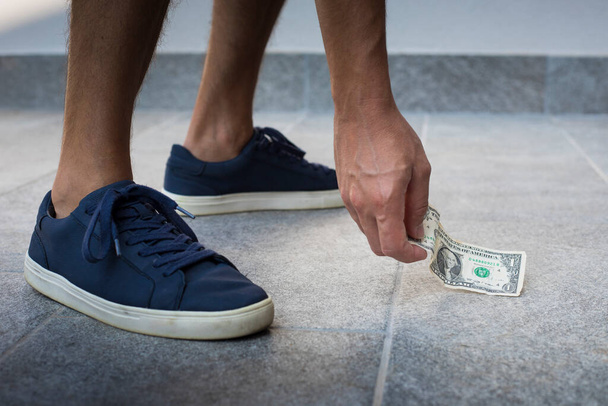 Anonymous man, casual dressed, with blue sneakers, picking up one dollar bill from the floor. - Foto, imagen