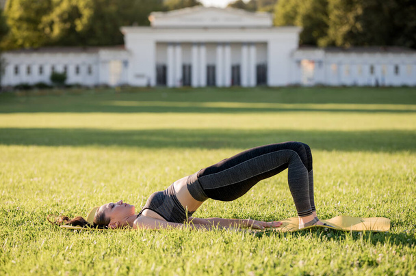 Mulher bonita deitada em um colchão amarelo, posar enquanto vestindo uma roupa de esportes apertados no parque fazendo pilates ou ioga, exercícios de iniciante de ponte de ombro - Foto, Imagem
