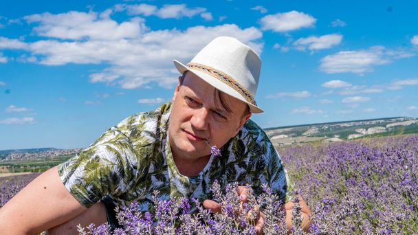 Smiling guy in lilac field looks seriously at the flowers sitting in a cap against the blue sky - Фото, изображение