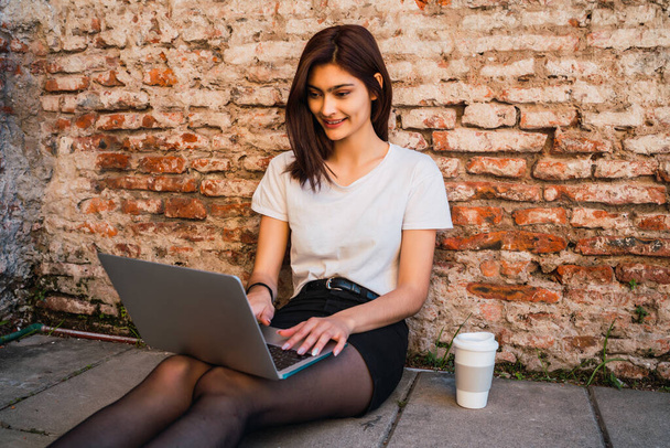 Portrait of young latin woman relax and using laptop while sitting outdoors against brick wall. Urban concept. - Fotó, kép