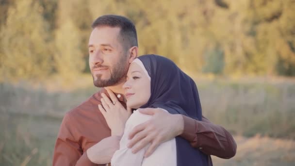 Feliz historia de amor musulmán de pareja mixta que abraza, acaricia y mira el atardecer sobre fondo verde. La mujer vestida con hijab mira a su hombre. Anuncio de agencia de citas on-line - Metraje, vídeo