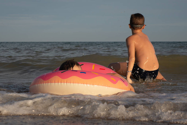 Братья 5 и 10 лет плавают и веселятся в море у берега. Младший мальчик с надувным кругом в форме пончика, старший в зеленых очках для плавания - Фото, изображение