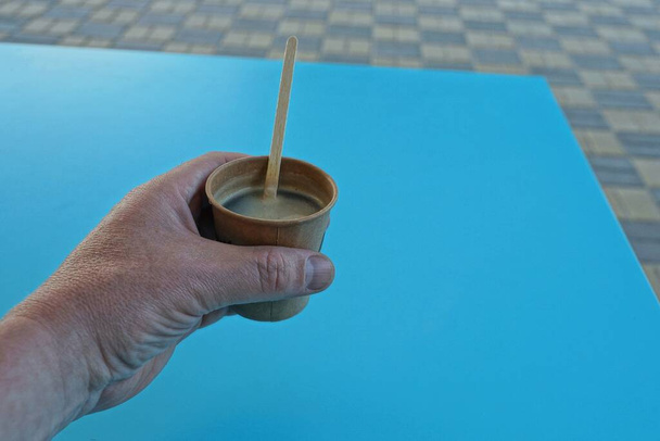 ruka drží hnědý papírový kelímek s kávou přes modrý stůl na chodníku na ulici - Fotografie, Obrázek