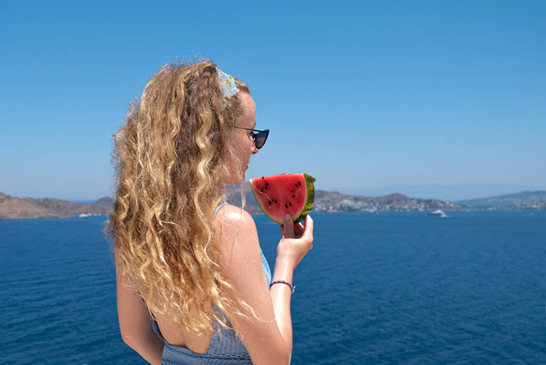 beautiful woman with a slice of watermelon wearing swimsuit eating red ripe watermelon slice with sea view holiday summer.back view. - Photo, Image