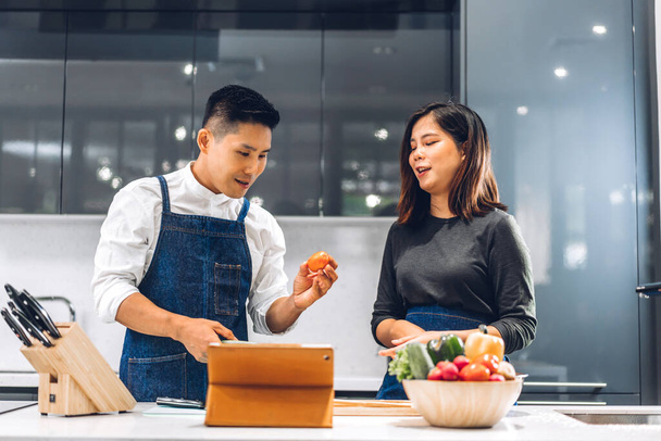 Genç Asyalı çift masada taze sebze salatasıyla yemek yaparken eğleniyor. Mutfakta lezzetli bir yemek hazırlamak için tablet bilgisayarlı ve internette yemek tarifi arayan mutlu çift. - Fotoğraf, Görsel