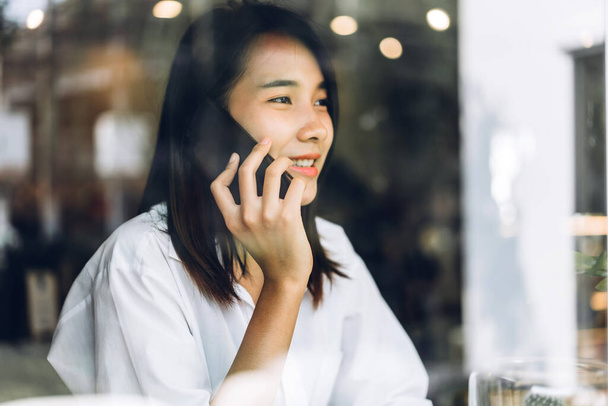 Retrato de sonriente feliz hermosa mujer asiática relajante usando smartphone.Young digital chica asiática hablar con un amigo en la cafetería - Foto, imagen