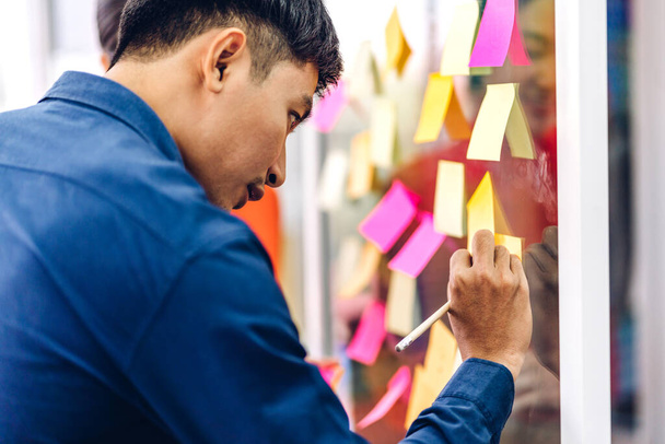 Casual creative happy asian businessman planning strategy analysis putting post it stickers note to share idea for startup project on glass board at modern office - Photo, Image