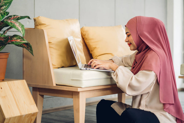 Genç, gülümseyen Müslüman kadın dizüstü bilgisayarını kullanarak rahatlıyor ve evde video konferansı yapıyor ev konseptinden çalışıyor. - Fotoğraf, Görsel
