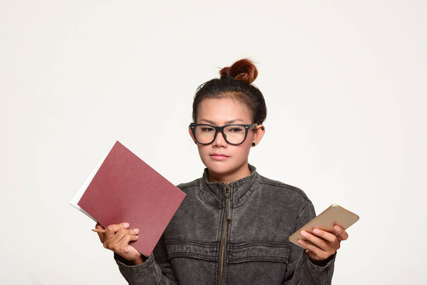 Asyalı bir kadın elinde bir kitap ve beyaz bir arka planda dikkatle telefonunu tutuyor.. - Fotoğraf, Görsel