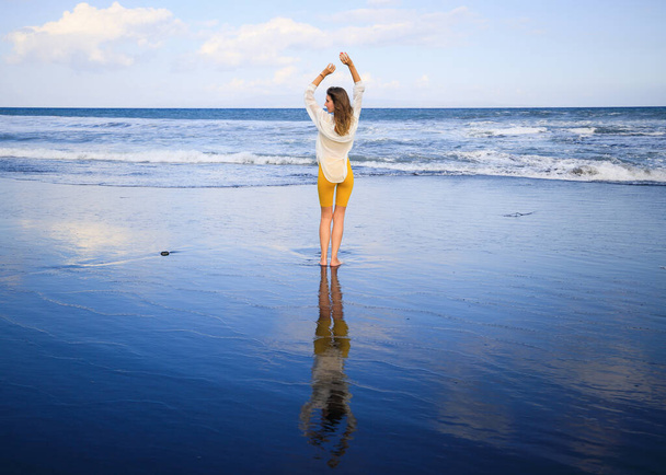 Krásná mladá žena kráčí po černé písečné pláži. Běloška ve žlutém sportovním oblečení a bílé blůze. Vodní odraz. Cestovní životní styl. Pohled zezadu. Rozumím. Bali, Indonésie - Fotografie, Obrázek