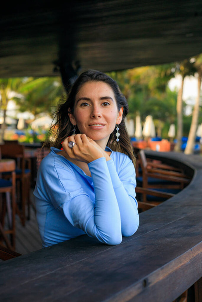 Primer plano retrato de mujer hermosa con joyas. Mujer caucásica sonriente con vestido azul y sentado en el bar de la playa. Estilo de vida. Disfruta la vida. Bali, Indonesia - Foto, imagen