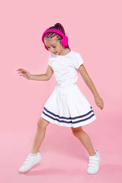 プリスクールの子供はピンクの背景でヘッドフォンで踊っています。スタジオで踊る幸せな女の子。全長像. - 写真・画像
