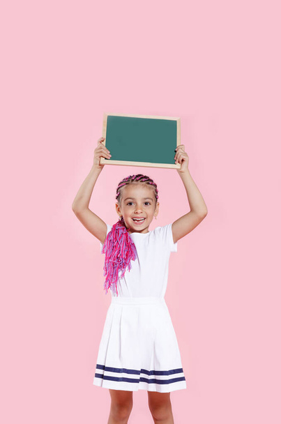 Smiling cute girl with pink ponytail, holding a small blackboard up, over pink background, standing in studio. Kid with empty chalkboard for your text, copy space. - Photo, Image