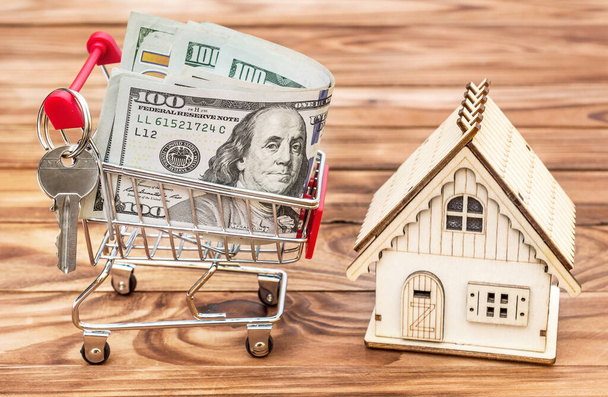 Nákupní košík s penězi a modelem domu na dřevěném pozadí. Koncept nákupu a prodeje nemovitostí. - Fotografie, Obrázek