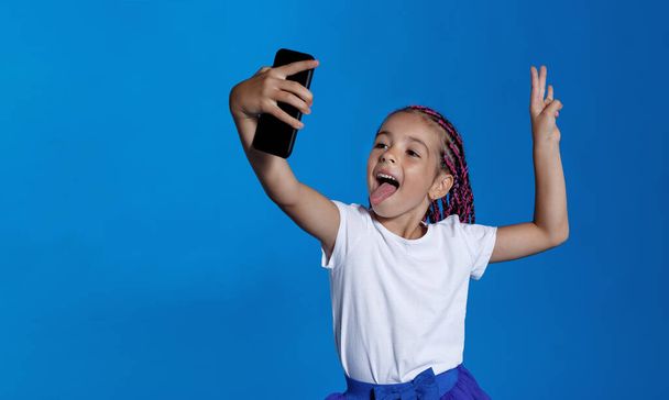 Horizontal view. Portrait of smiling amazing little girl holding in hand a smartphone doing selfie over blue background. Space for text. - Zdjęcie, obraz
