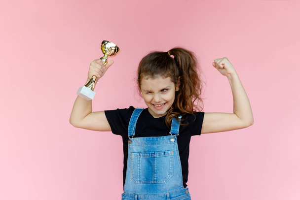 Portrait of a happy little girl, in denim clothes, smiling, with closed eyes, showing a successful gesture of a winner, holding in hand a prize, over pink background. Copy space. Horizontal view. - Photo, Image