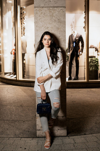 Atraktivní žena v bílé módní bundě a džínách se dívá do kamery. Mladá žena na pozadí budovy - Fotografie, Obrázek