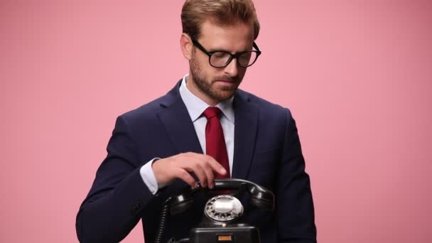 sorridente jovem empresário vestindo óculos atender telefone, falando, acenando e verificando o tempo, concordando e rindo em fundo rosa - Filmagem, Vídeo