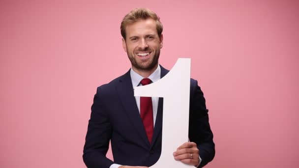 smiling young fashion model in navy blue suit holding number one sign and making thumbs up on pink backgrounds - Footage, Video