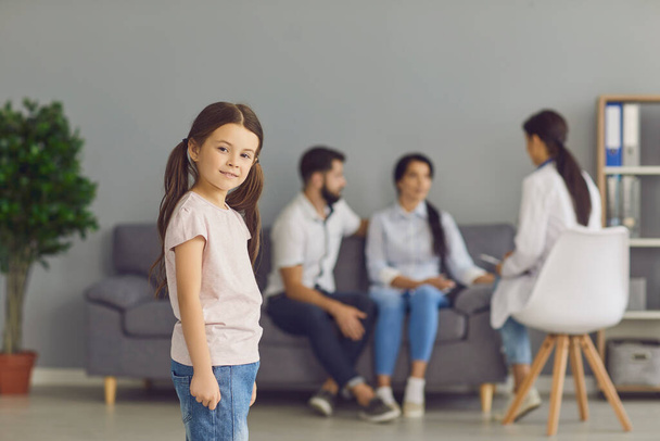 Smiling girl standing in medical clinic over parents talking to woman doctor at background - 写真・画像