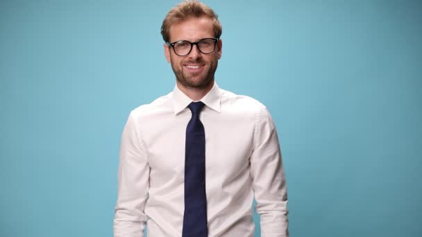 seksowny biznesmen z okularami i uśmiechem na twarzy, trzymający jedną rękę w kieszeni i celujący w kamerę na niebieskim tle - Materiał filmowy, wideo